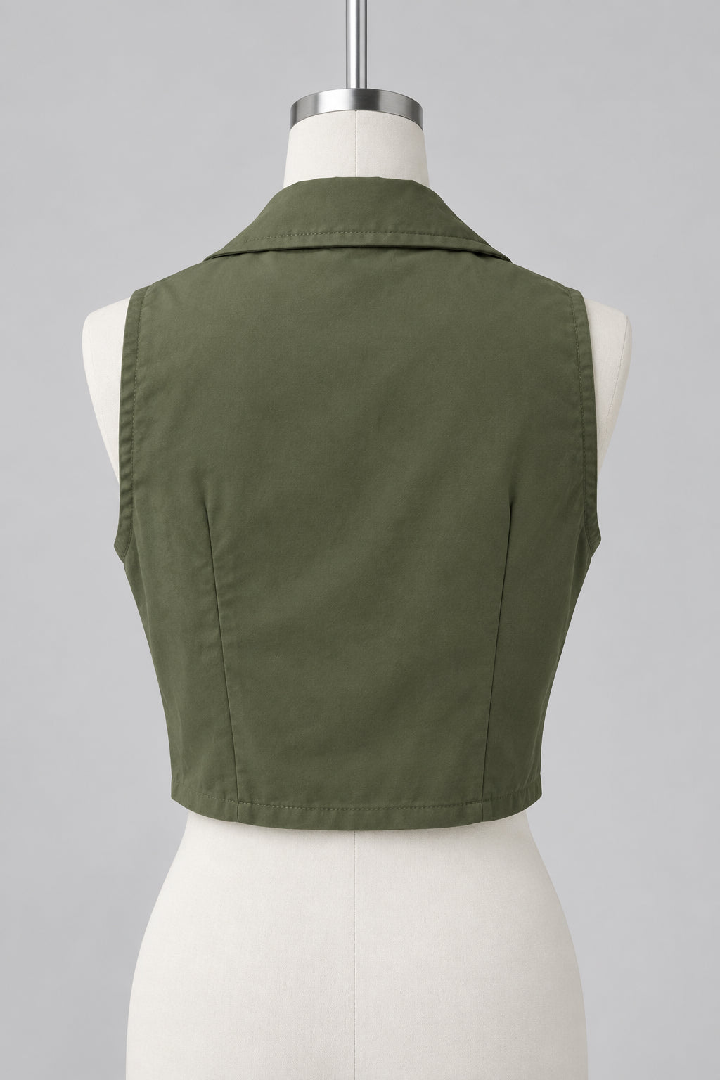 An olive green sleeveless collared vest displayed on a white dress form mannequin, photographed in an indoor room with windows, curtains, and a plant visible in the background.