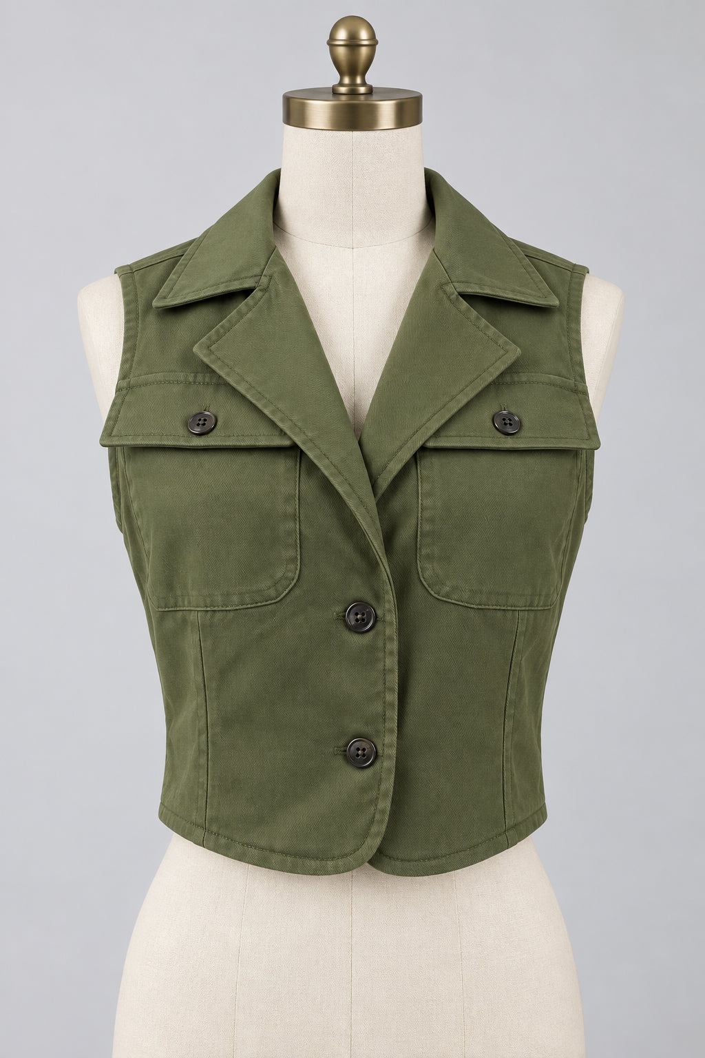 An olive green cropped utility vest displayed on a white mannequin torso. The vest features a pointed collar, three dark metal snap buttons down the front, and two flap pockets on the chest.