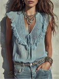 A woman wearing a light blue sleeveless blouse with ruffle trim and a tie-front detail, paired with distressed blue jeans and layered necklaces, photographed against a textured light wall.