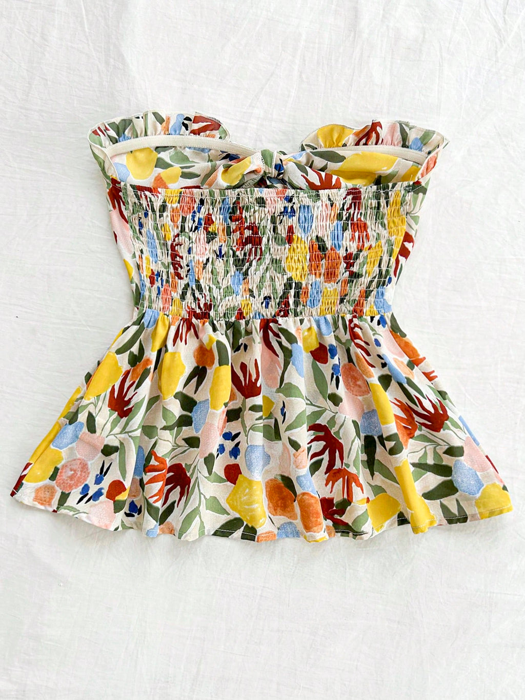 A sleeveless smocked top with a ruffled peplum, featuring a vibrant multicolor floral and botanical print on a cream background, laid flat on a white fabric backdrop.