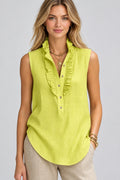 A woman wearing a sleeveless ruffled blouse in a bright yellow-green color, paired with beige pants and gold jewelry, set against a blurred outdoor background.