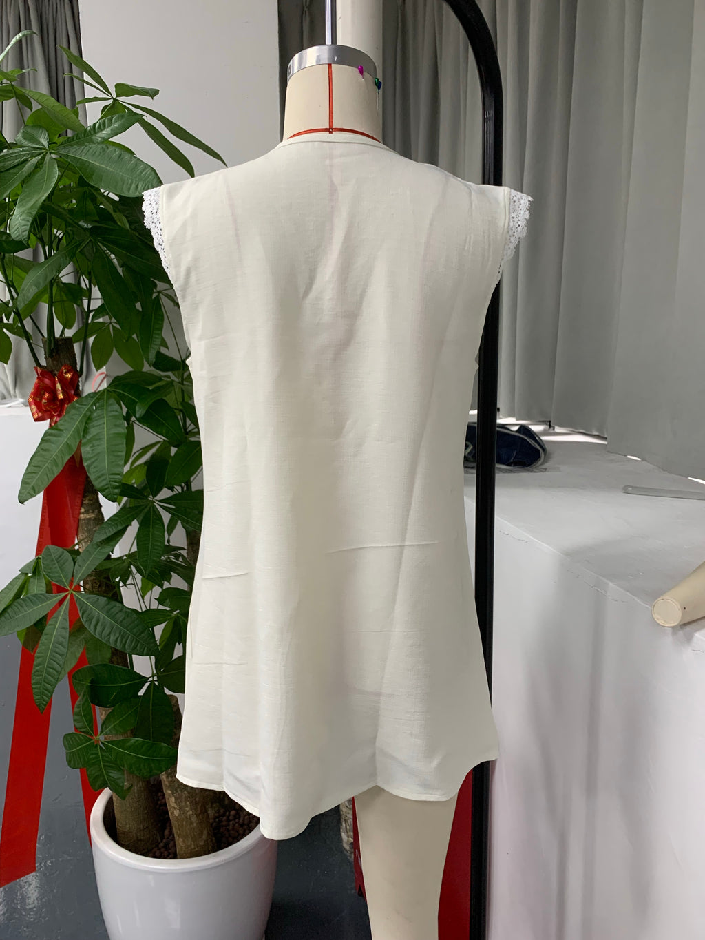 A sleeveless white tunic top with delicate lace trim at the shoulders, displayed on a dress form mannequin in an indoor setting with a potted plant and table in the background.