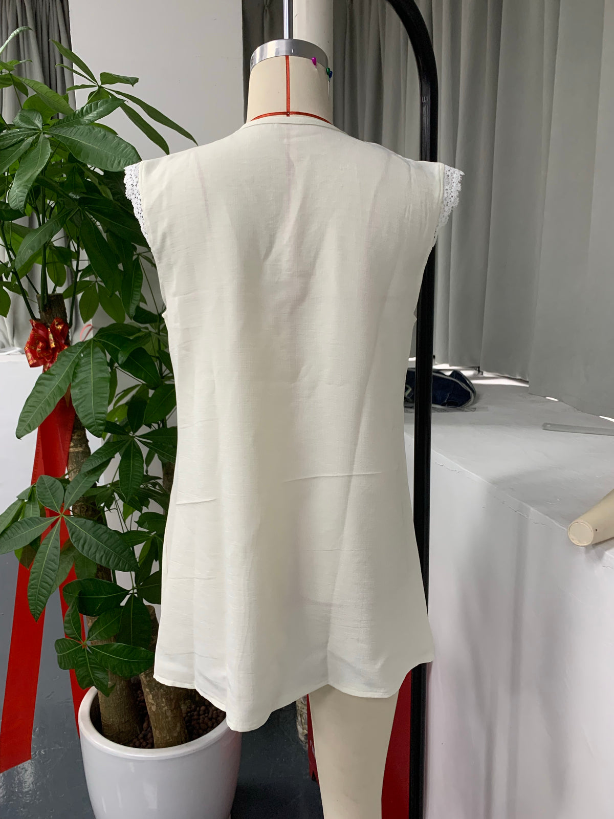 A sleeveless white tunic top with delicate lace trim at the shoulders, displayed on a dress form mannequin in an indoor setting with a potted plant and table in the background.