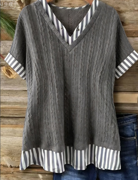 A gray short-sleeve V-neck top with a cable-knit center panel and striped trim on the collar, sleeve cuffs, and hem, shown hanging against a rustic wood plank background.
