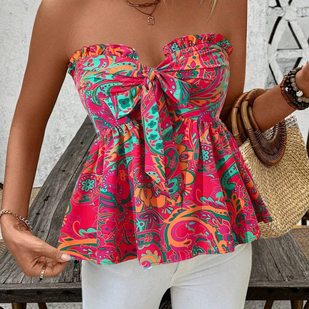 A woman wearing a strapless ruffled peplum top with a large front bow, featuring a bright paisley pattern in pink, teal, orange, and yellow. She is also wearing white pants and holding a woven straw bag.