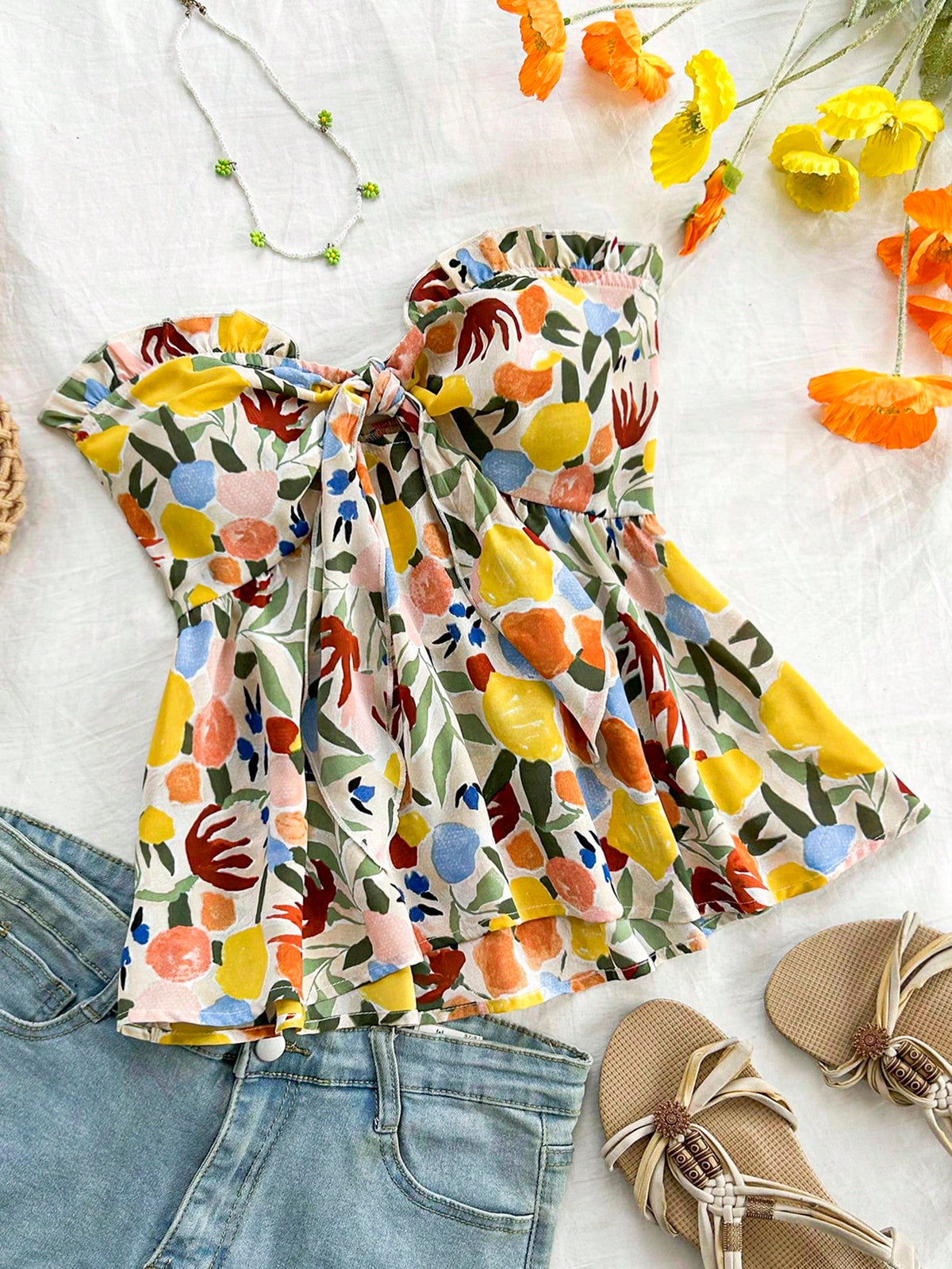 A colorful floral print strapless top with a ruffled sweetheart neckline, styled in a flat lay with blue jeans, beige sandals, and flowers on a white fabric background.